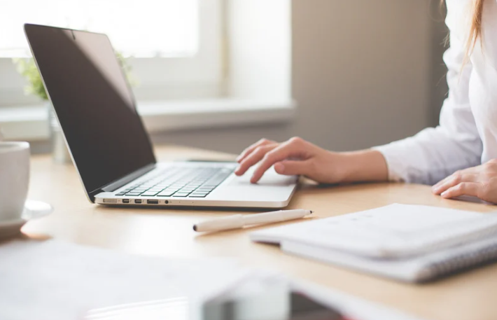 Nahaufnahme einer Hand, die auf einem Laptop tippt. Der Bildschirm zeigt eine geöffnete Arbeitsdatei. Der Arbeitsplatz ist modern und gut organisiert, mit einem Notizblock und einer Tasse Kaffee neben dem Laptop.